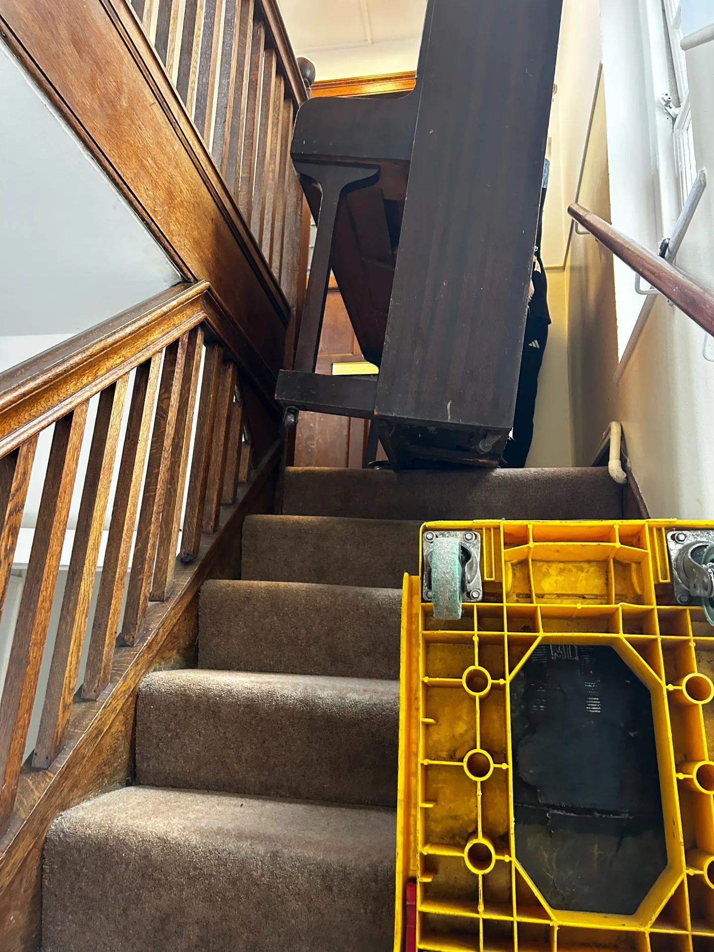 Upright piano being moved on a carpeted staircase, seen from the bottom of the stairs